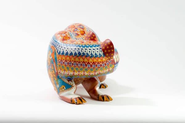 bear mexican wood carving alebrije This incredible bear carving by Ray Medez has intricate painting featuring pre-hispanic iconography and symbolism. Each of the four legs has an illustration. The front left leg shows the Aztec God Quetzalcoatl (represented by the feathered snake). The back right leg shows Kukulcán, the Mayan wisdom deity related to Quetzalcoatl.The front right leg depicts the Water-Lily Jaguar, an anthropomorphic form of an important Maya god. His special attribute is a water-lily atop his head, which may reflect the jaguar’s love of water. This jaguar god moves and acts like a human. His necklace is called a death-eye collar and is part of his attire for the sacrificial death dance. The Water-Lily Jaguar is a significant character in Maya narrative artwork.The scene on the back left leg represents the extraction of the spirit beverage pulque from the maguey plant. The seashells on the body represent fertility and birth. It is also a symbol related to Quetzalcoatl.Extraordinary Mexican art from MexArt, London UK.