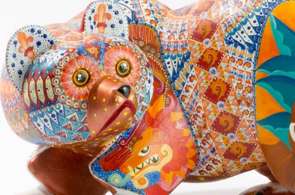 bear mexican wood carving alebrije This incredible bear carving by Ray Medez has intricate painting featuring pre-hispanic iconography and symbolism. Each of the four legs has an illustration. 
The front left leg shows the Aztec God Quetzalcoatl (represented by the feathered snake). 
The back right leg shows Kukulcán, the Mayan wisdom deity related to Quetzalcoatl.
The front right leg depicts the Water-Lily Jaguar, an anthropomorphic form of an important Maya god.  His special attribute is a water-lily atop his head, which may reflect the jaguar’s love of water.  This jaguar god moves and acts like a human.  His necklace is called a death-eye collar and is part of his attire for the sacrificial death dance.  The Water-Lily Jaguar is a significant character in Maya narrative artwork.
The scene on the back left leg represents the extraction of the spirit beverage pulque from the maguey plant. 
The seashells on the body represent fertility and birth. It is also a symbol related to Quetzalcoatl.
Extraordinary Mexican art from MexArt, London UK.